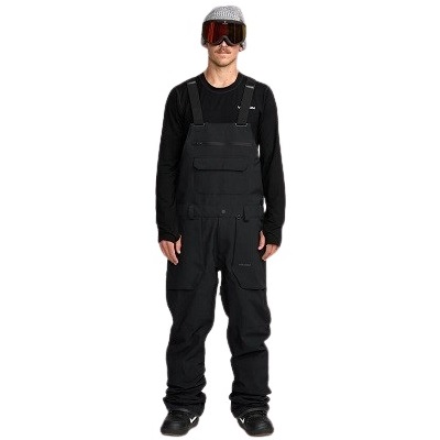 Man wearing black bib overalls with chest pocket, black long-sleeve shirt, ski goggles, and a patterned beanie, standing against a white background.