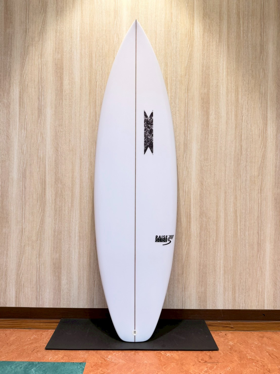 White surfboard standing upright on a black mat against a wood-paneled wall, with a small black logo near the bottom.