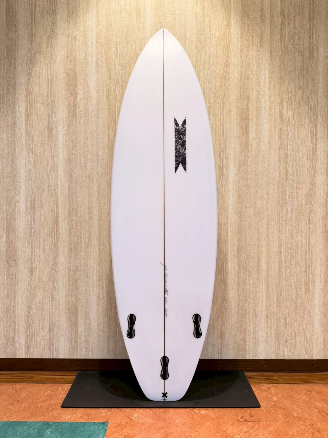 White surfboard standing upright on a black display stand against a wood-paneled wall; fin boxes visible along the rails, minimal branding near center.