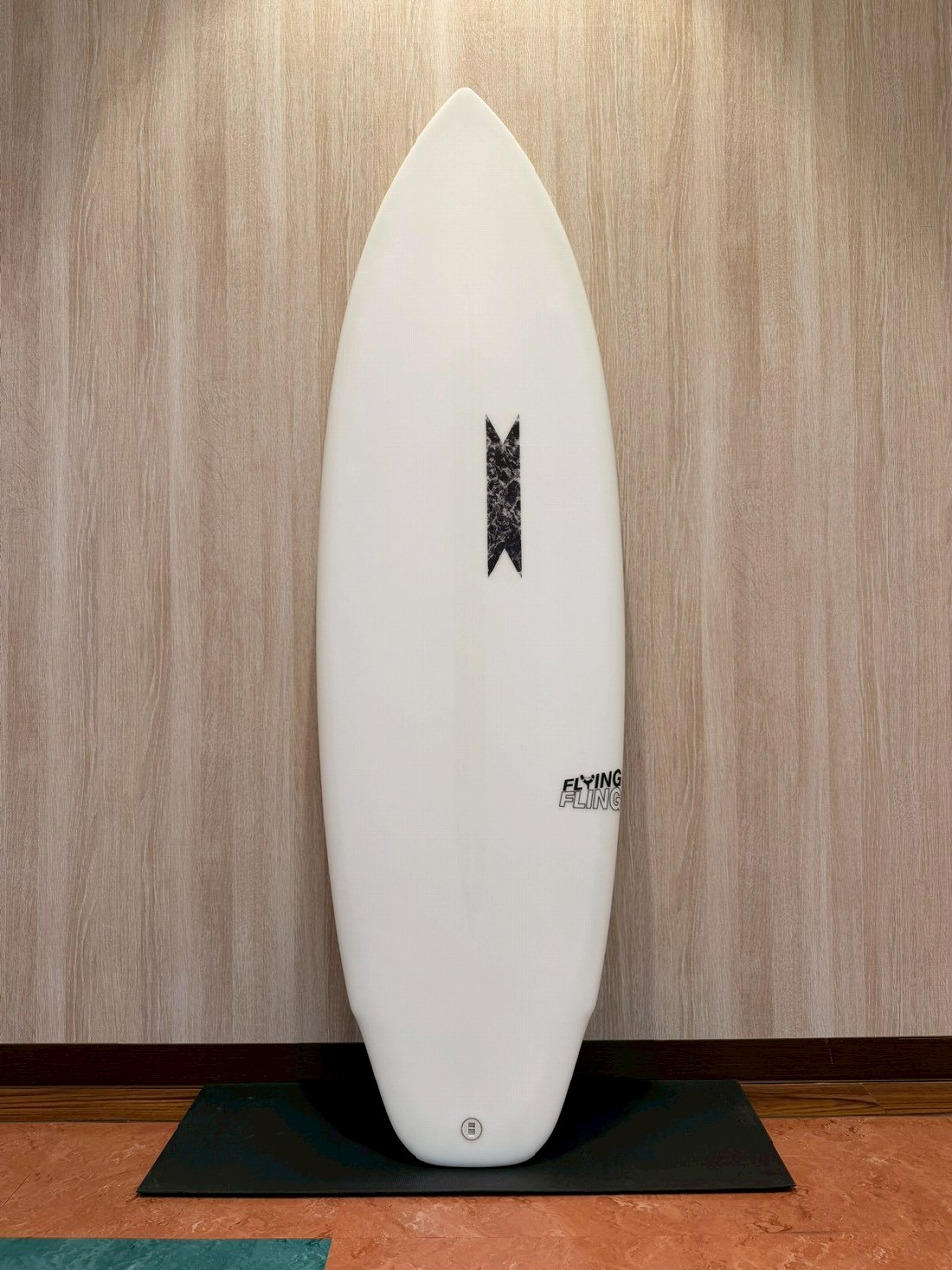 White surfboard standing upright on a black mat against a wood-paneled wall, with a small black central logo and a round tail plug.
