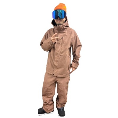 Person wearing a brown waterproof snowsuit with hood, orange beanie and blue goggles, standing with a hand in pocket.