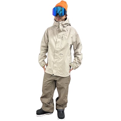 Person wearing a beige winter jacket, khaki snow pants, orange beanie, and blue ski goggles standing with hands in pockets.