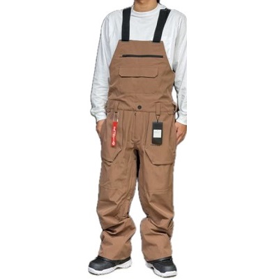 Person wearing tan bib overalls with black suspenders over a white long-sleeve shirt, standing with hands in pockets.