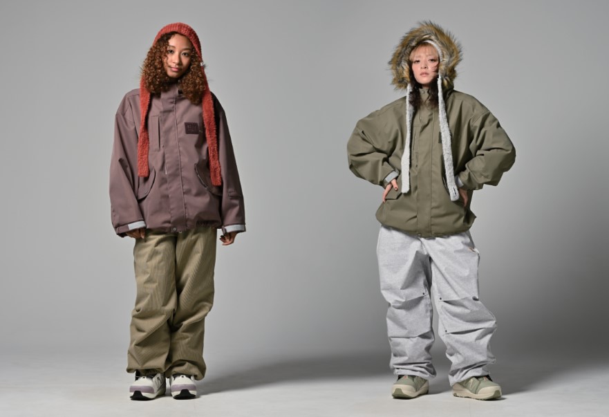 Two models stand in a studio wearing oversized winter jackets and baggy pants; left in mauve jacket with red knit hood, right in olive jacket with fur-lined hood.