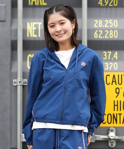 Smiling woman in a blue zip-up windbreaker over a white shirt, standing in front of a metal shipping container with yellow numbers.