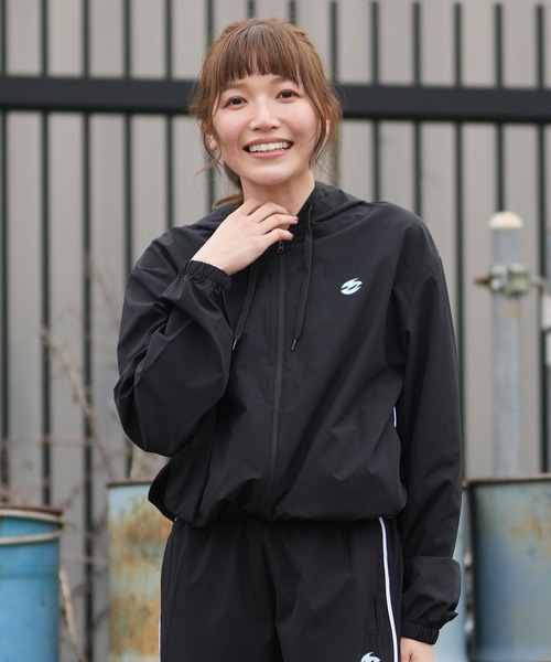 Woman smiling outdoors in a black windbreaker and matching pants, with a metal fence behind her.