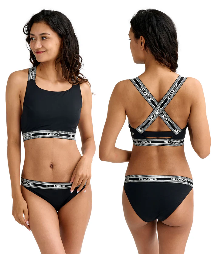 Woman modeling a black sporty bralette and matching bikini briefs with logo waistband; front view with hands at sides and back view showing crossed straps and logo elastic.