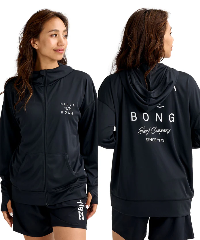 Two women wear black surf jackets and shorts, one facing forward and the other showing the back with a large Billabong logo and 'Since 1973' text.