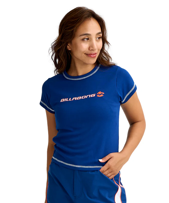 Woman in a blue Billabong t-shirt with white stitching, looking to her right, posed in a casual athletic outfit.