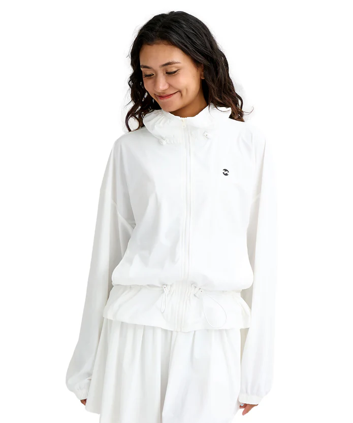 Woman wearing a white zip-up windbreaker with a drawstring waist and matching pants, small black logo on the chest, smiling downward.