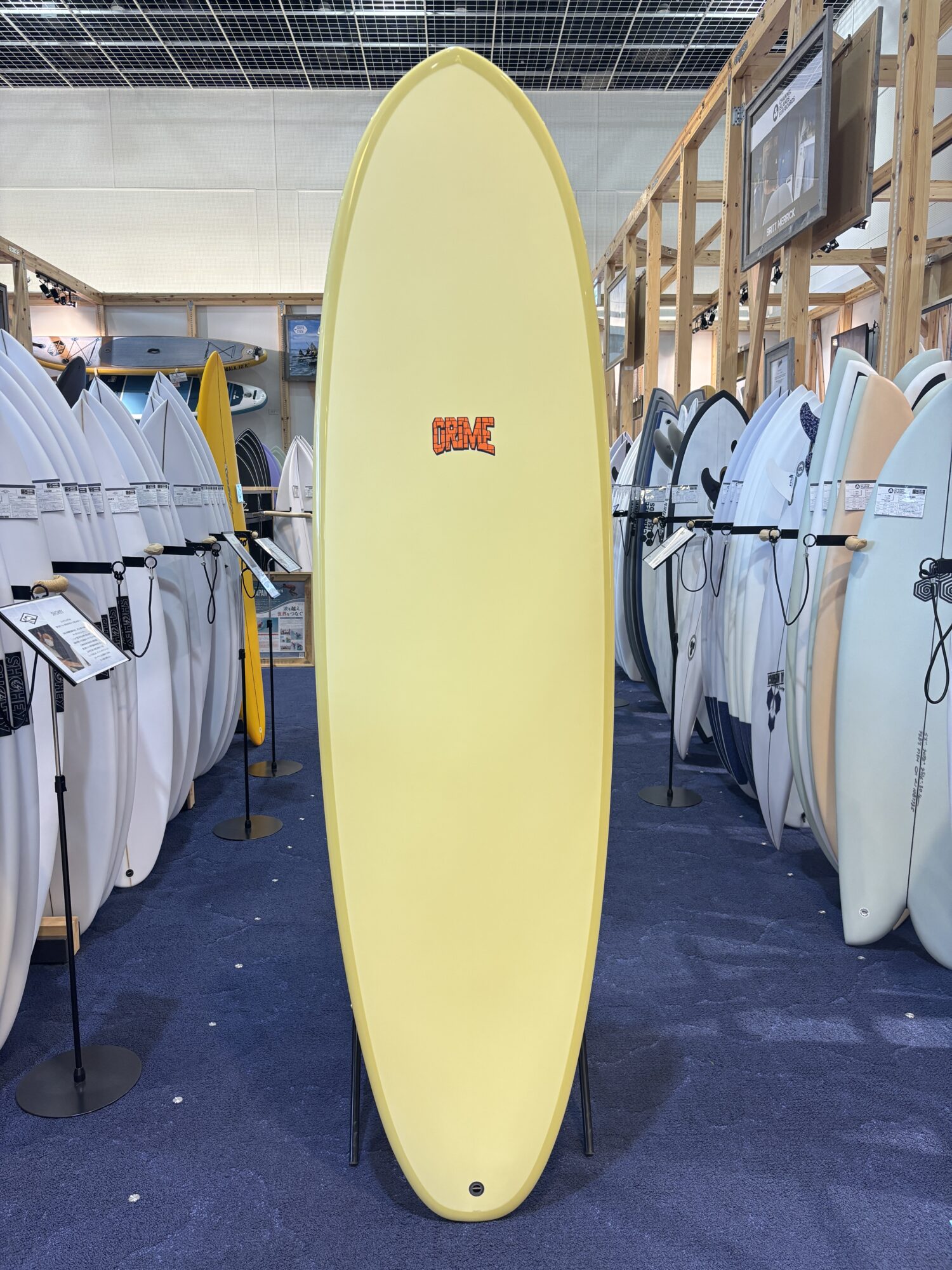 IMG_3950 - SHOP NEWS | ムラサキスポーツオンラインストア 通販 Yellow surfboard on display in a surf shop, surrounded by racks of white and colored boards on both sides.