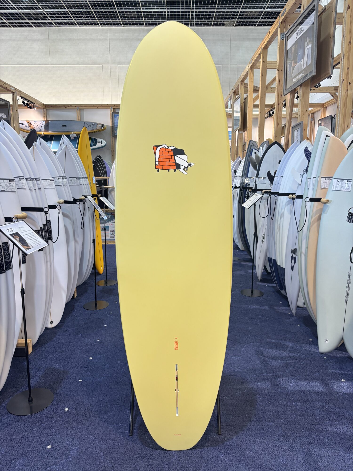 IMG_3951 - SHOP NEWS | ムラサキスポーツオンラインストア 通販 Yellow surfboard on display in a showroom, surrounded by rows of other boards.