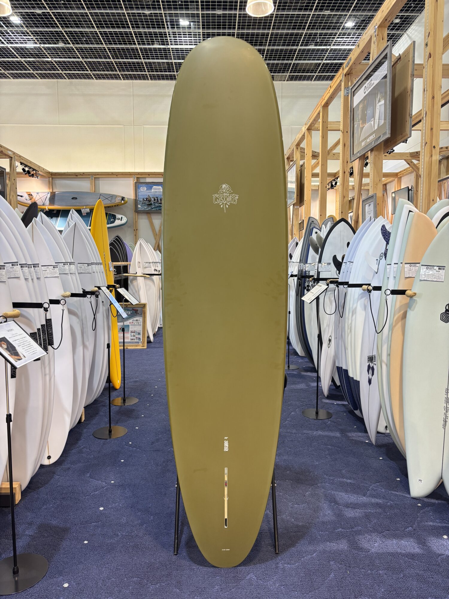 IMG_3954 - SHOP NEWS | ムラサキスポーツオンラインストア 通販 Olive-green surfboard standing upright on a display rack in a showroom surrounded by other surfboards.