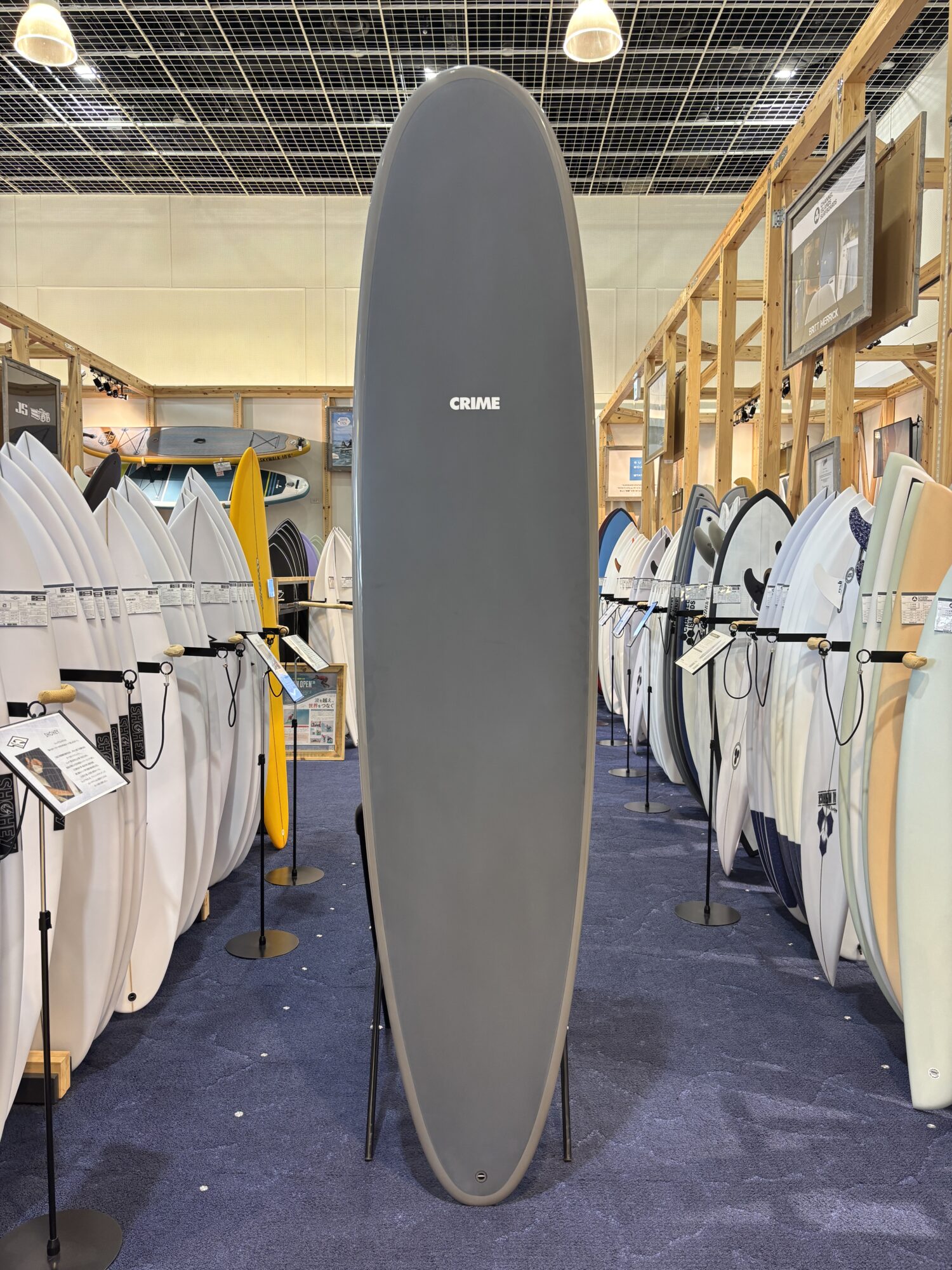IMG_3955 - SHOP NEWS | ムラサキスポーツオンラインストア 通販 Tall gray surfboard standing upright in a showroom aisle, surrounded by rows of white and colored boards on racks under a grid ceiling.