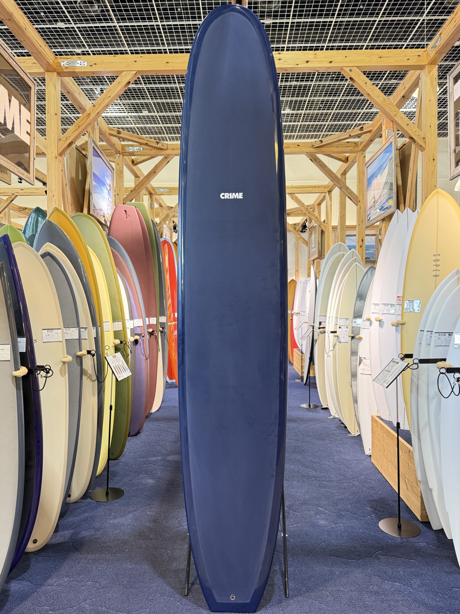 IMG_4061 - SHOP NEWS | ムラサキスポーツオンラインストア 通販 Tall blue surfboard standing upright in a showroom, surrounded by colorful surfboards on racks.