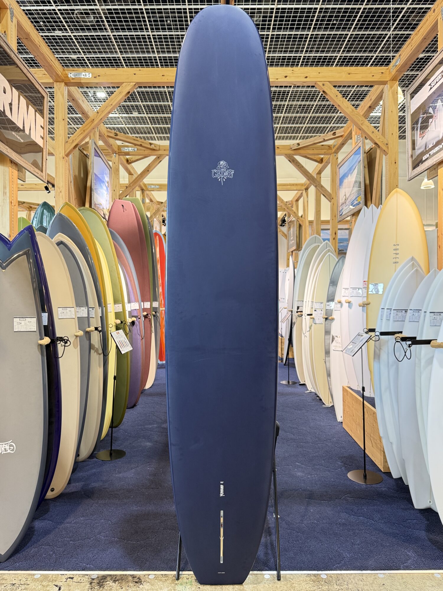 IMG_4062 - SHOP NEWS | ムラサキスポーツオンラインストア 通販 Tall blue surfboard standing upright on a display stand in a shop, surrounded by colorful boards on racks.