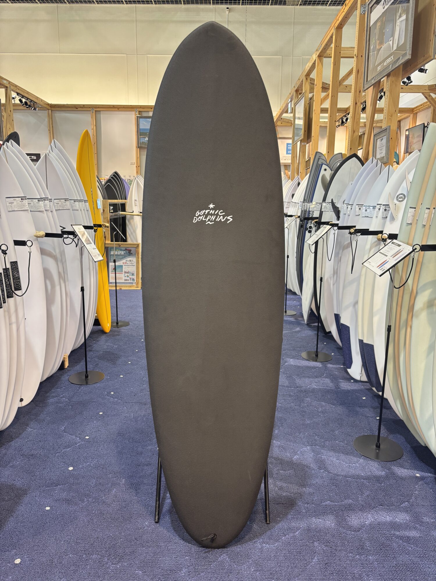 Tall black surfboard with Gothic Dolphins logo displayed upright in a showroom, surrounded by rows of white and colored boards on racks.