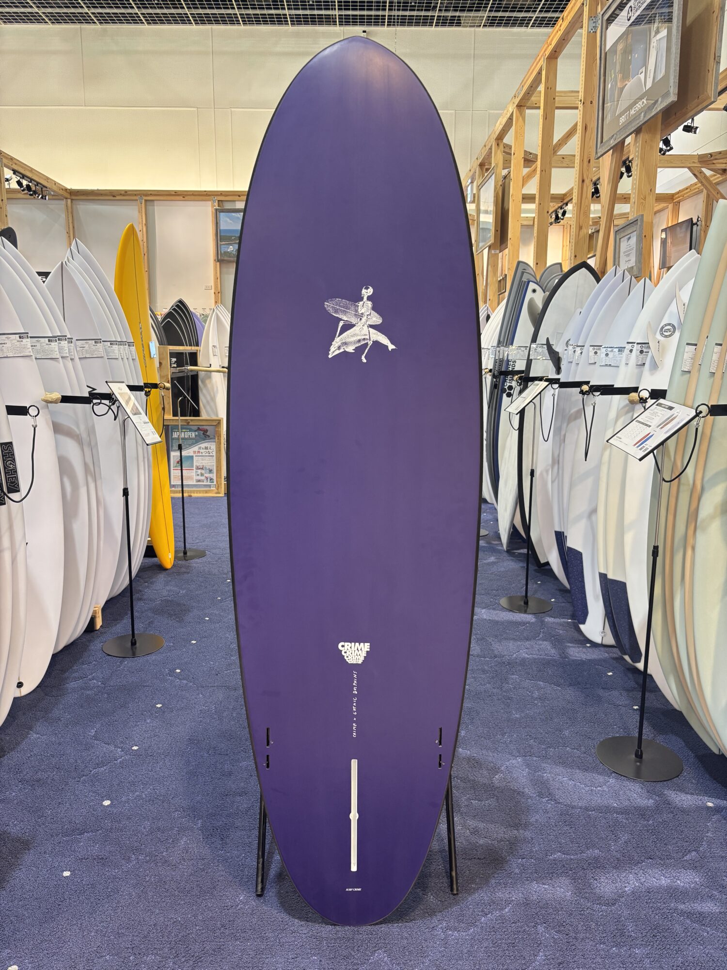 Tall purple surfboard on a stand in a showroom, surrounded by white and colored surfboards on racks.