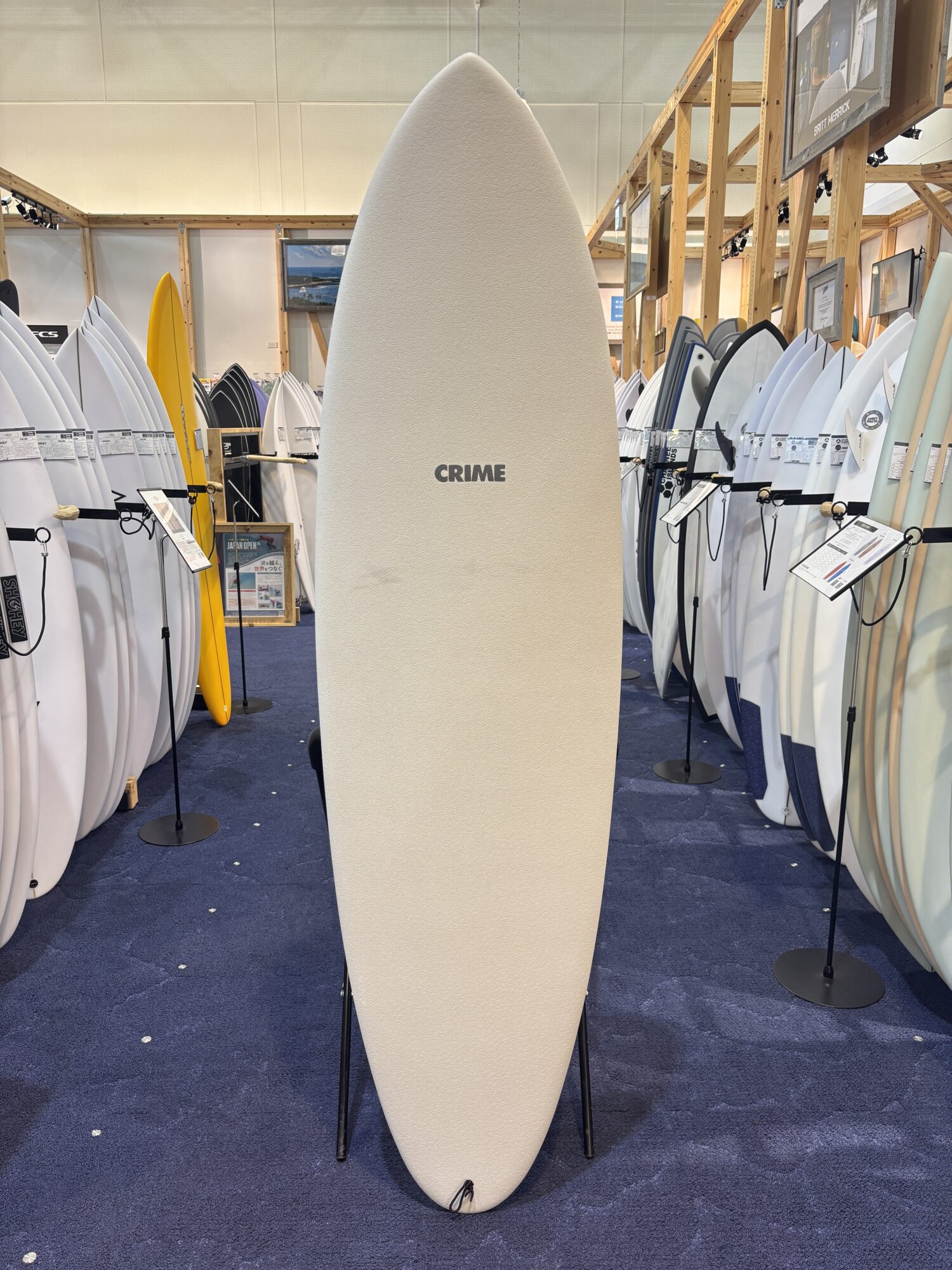 Large beige surfboard stands upright in a showroom, surrounded by rows of other surfboards on racks; the center board has 'CRIME' written on it.