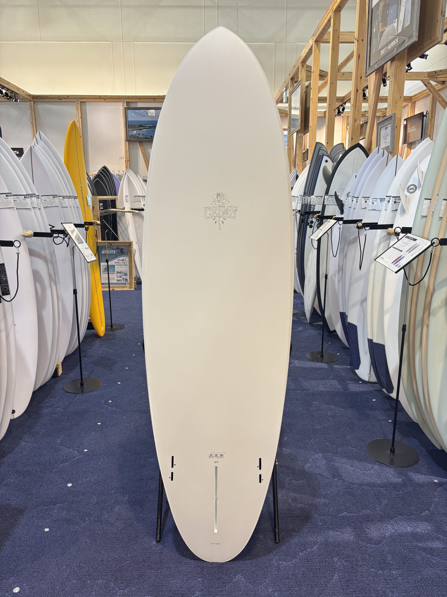 Tall white surfboard displayed upright on a stand in a showroom, surrounded by many other surfboards of various colors and sizes on racks.
