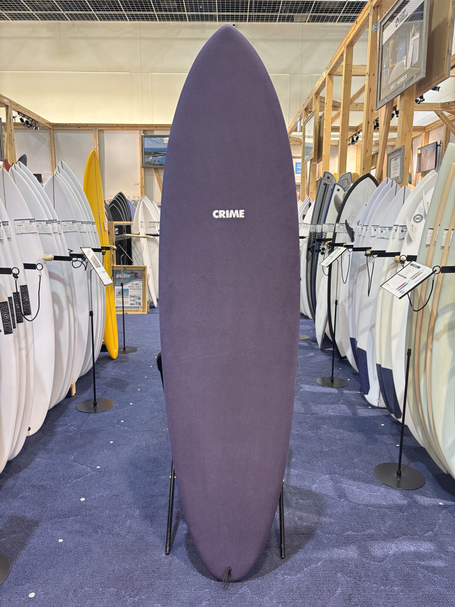Tall purple surfboard displayed upright in a showroom, flanked by white and colored boards on racks.