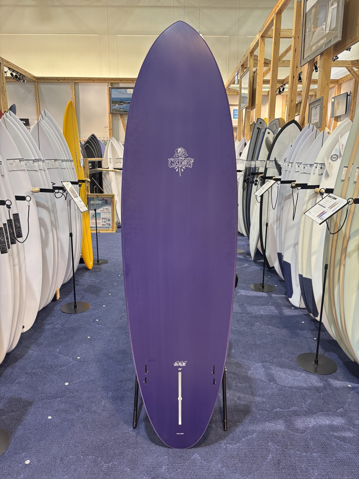 Tall purple surfboard standing upright in a showroom with white and colored boards lined up along wooden racks.