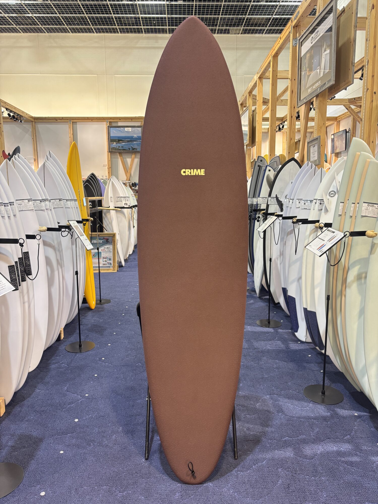 Tall brown surfboard with the word CRIME in yellow letters, standing upright among display boards in a surf shop showroom.