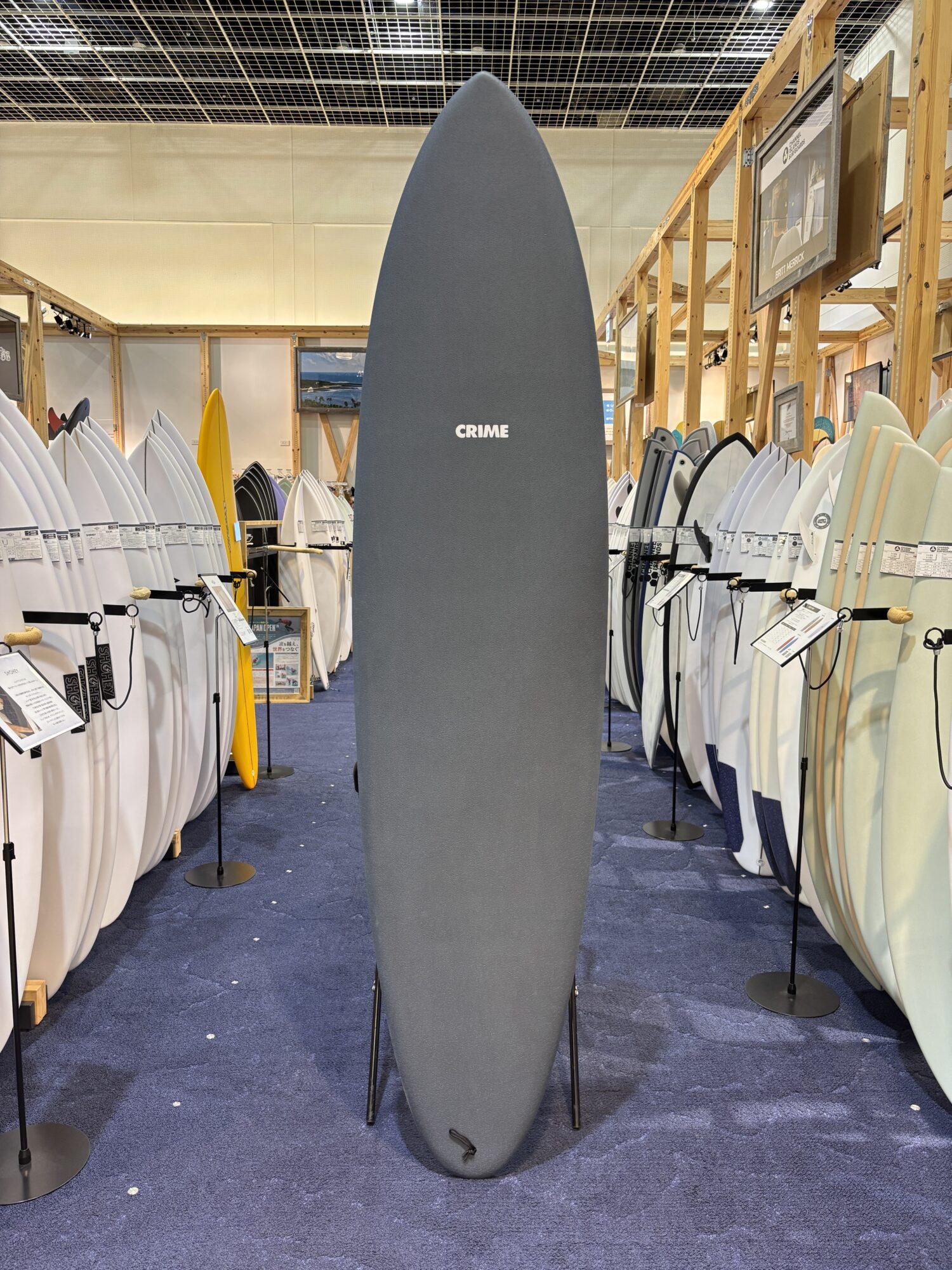 Tall, dark gray surfboard on display in a showroom, surrounded by many white and colored boards on racks.