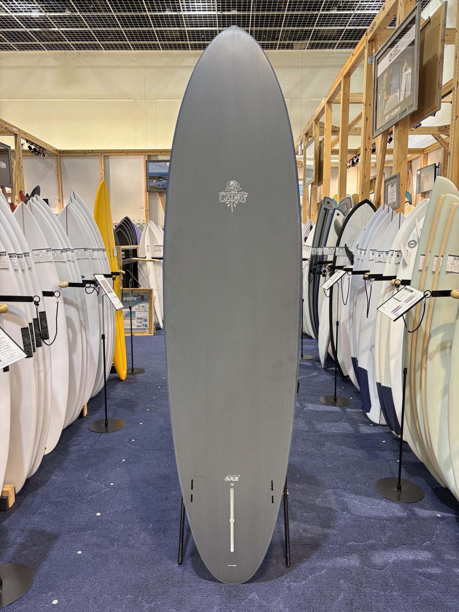 Tall gray surfboard on display in a shop, surrounded by many white and colored boards on racks.