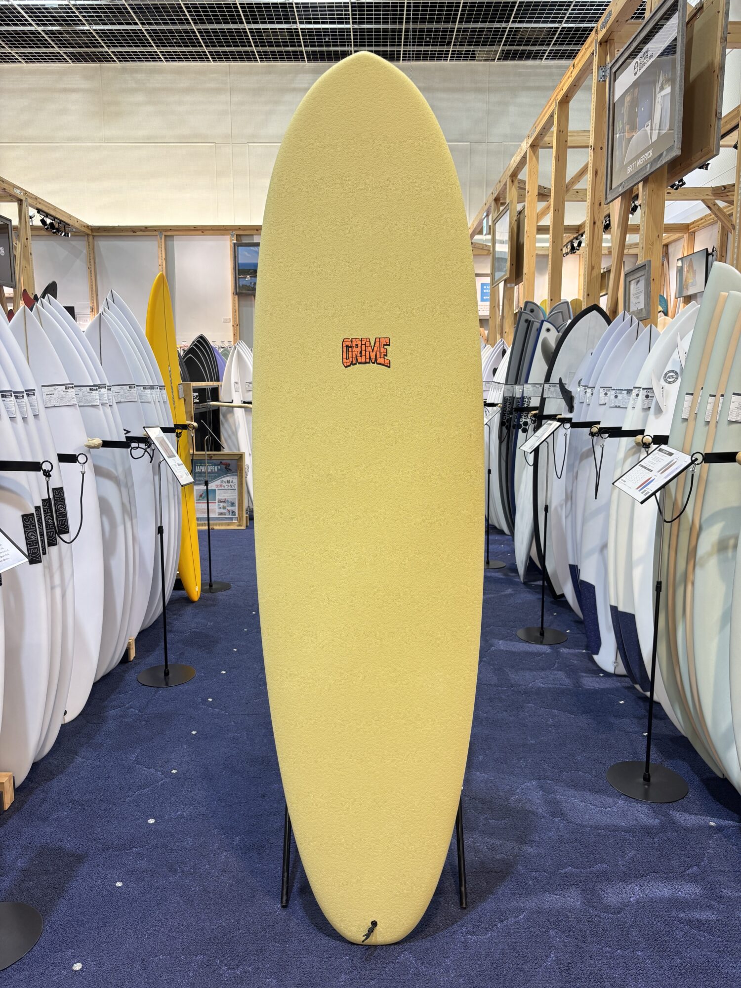 IMG_4183 - SHOP NEWS | ムラサキスポーツオンラインストア 通販 Tall yellow surfboard standing upright on a stand in a showroom, surrounded by rows of surfboards on both sides.