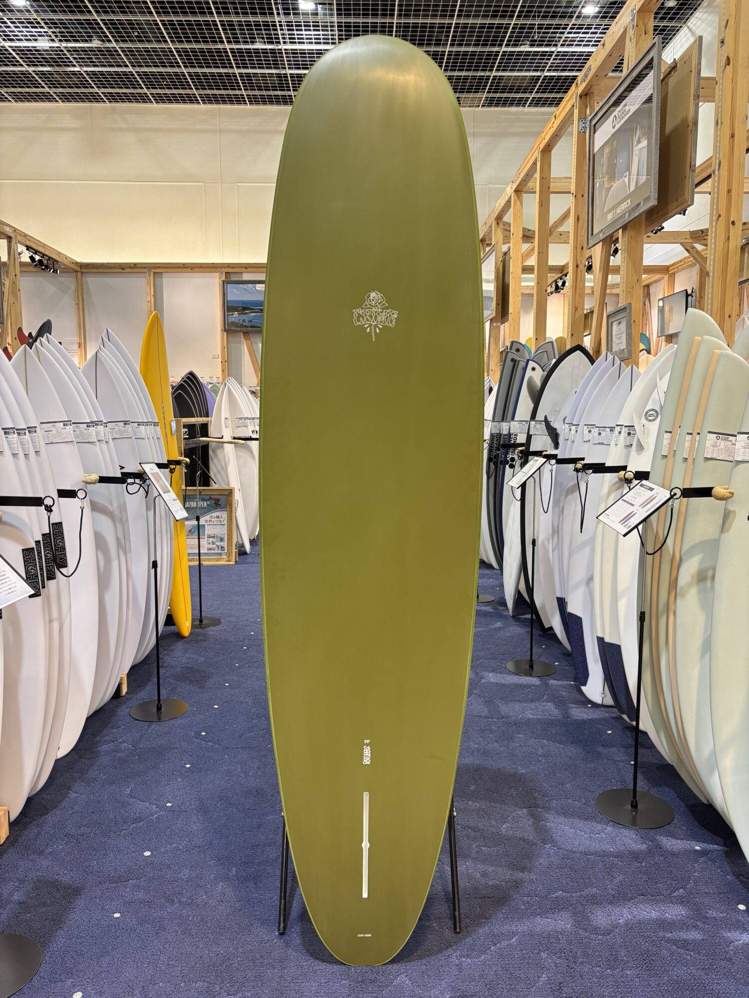 IMG_4186 - SHOP NEWS | ムラサキスポーツオンラインストア 通販 Olive-green surfboard on display in a showroom, surrounded by white and colorful boards on wooden racks.