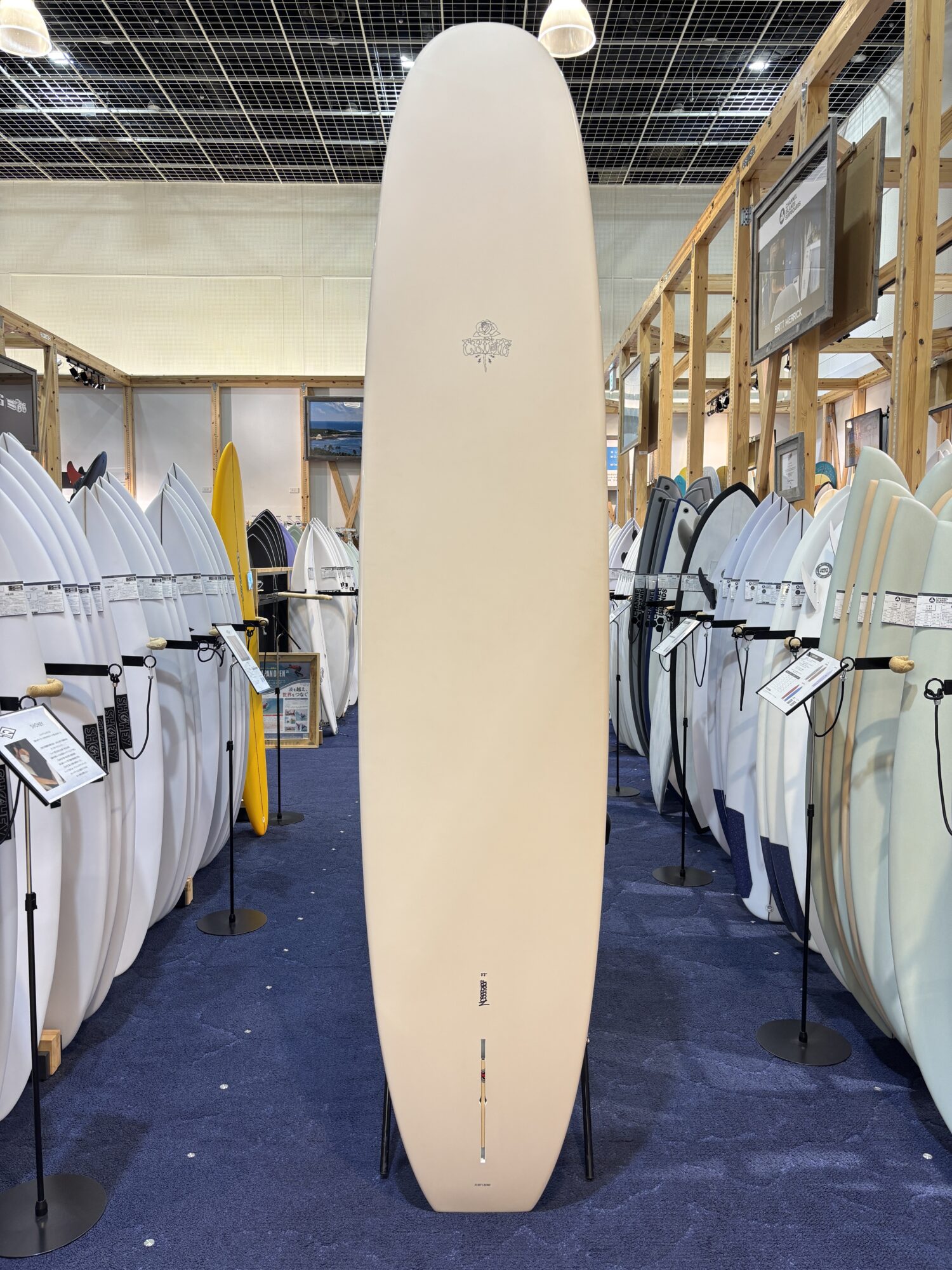 IMG_4188 - SHOP NEWS | ムラサキスポーツオンラインストア 通販 Tall beige surfboard on display stand in a showroom surrounded by rows of white and colored boards on racks.