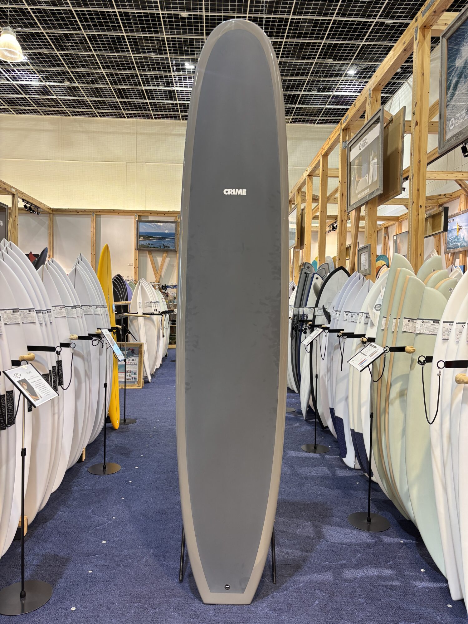 IMG_4189 - SHOP NEWS | ムラサキスポーツオンラインストア 通販 Tall gray surfboard with a small 'CRIME' logo stands in a showroom aisle, surrounded by rows of white and colored boards on racks.