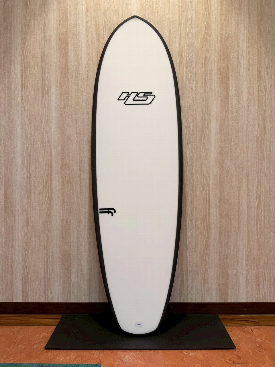 LINE_ALBUM_2026 ヘイデン_260524_1 - SHOP NEWS | ムラサキスポーツオンラインストア 通販 White surfboard with black edging standing upright against a wooden wall on a black display mat.
