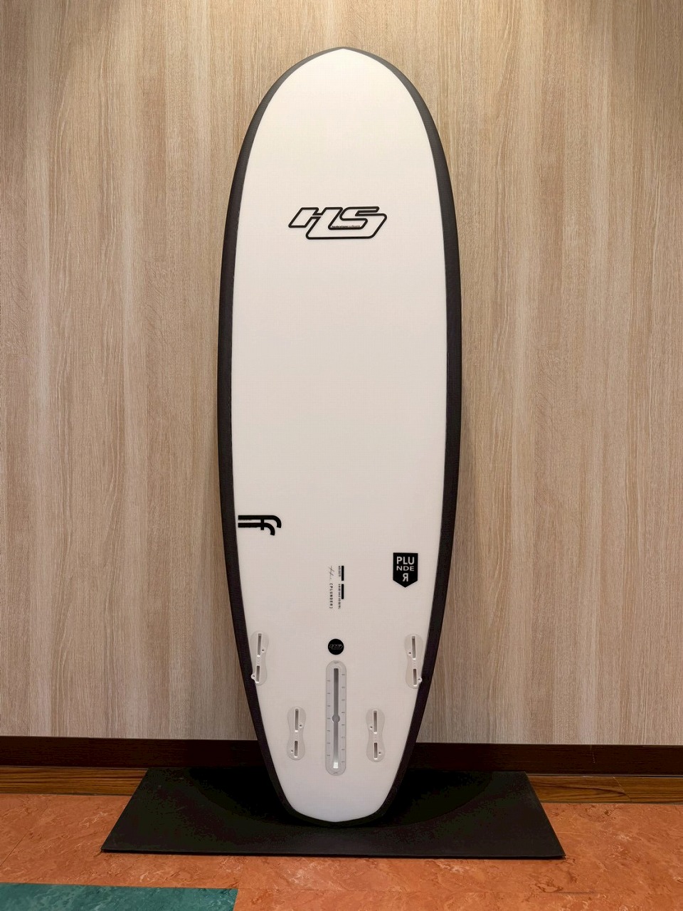 LINE_ALBUM_2026 ヘイデン_260524_2 - SHOP NEWS | ムラサキスポーツオンラインストア 通販 White surfboard with black edge trim, standing upright on a black mat against a wood-paneled wall; fin boxes visible near the tail.