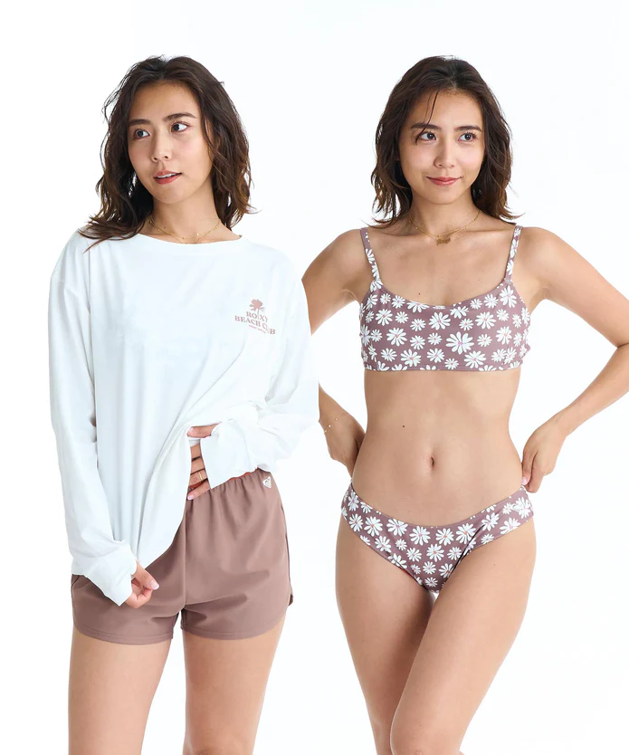 Two women stand side by side in summer outfits: one in a white long-sleeve tee and brown shorts, the other in a purple floral bikini set, against a white background.
