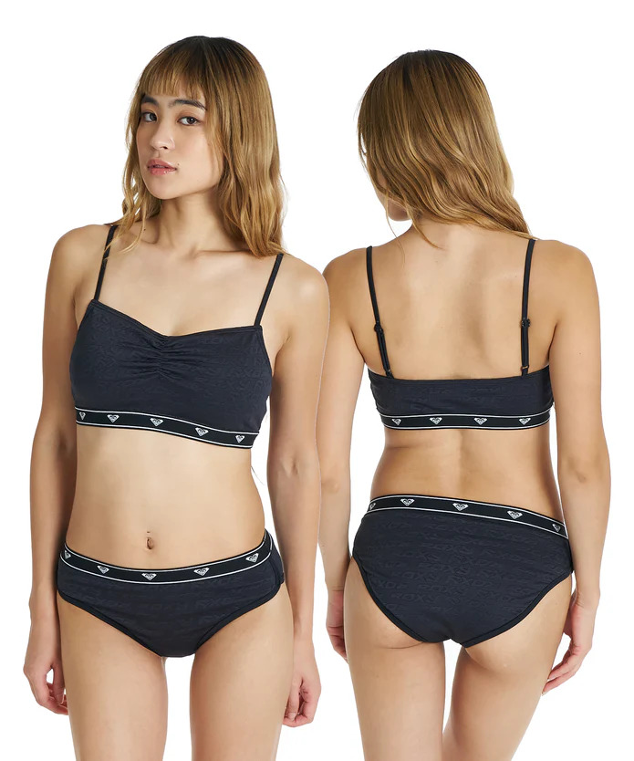 Model wearing black bralette and matching briefs with logo-pattern elastic bands, shown from the front and back.