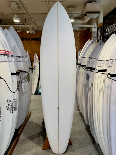 White surfboard displayed upright on a stand in a surf shop, with rows of other boards on either side.