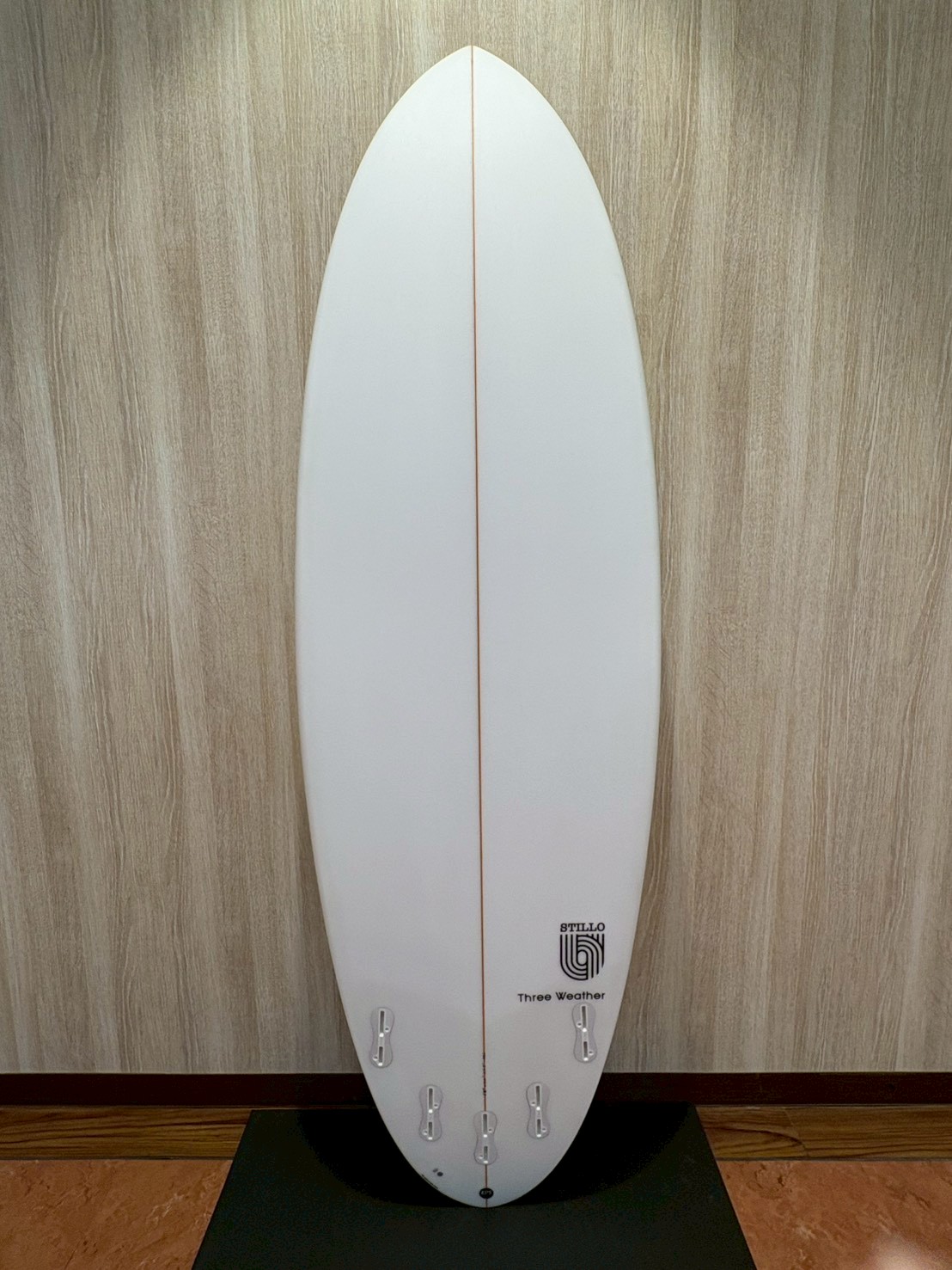 White surfboard standing upright indoors against a wood-paneled wall, with a thin center stringer and multiple fin boxes near the tail.
