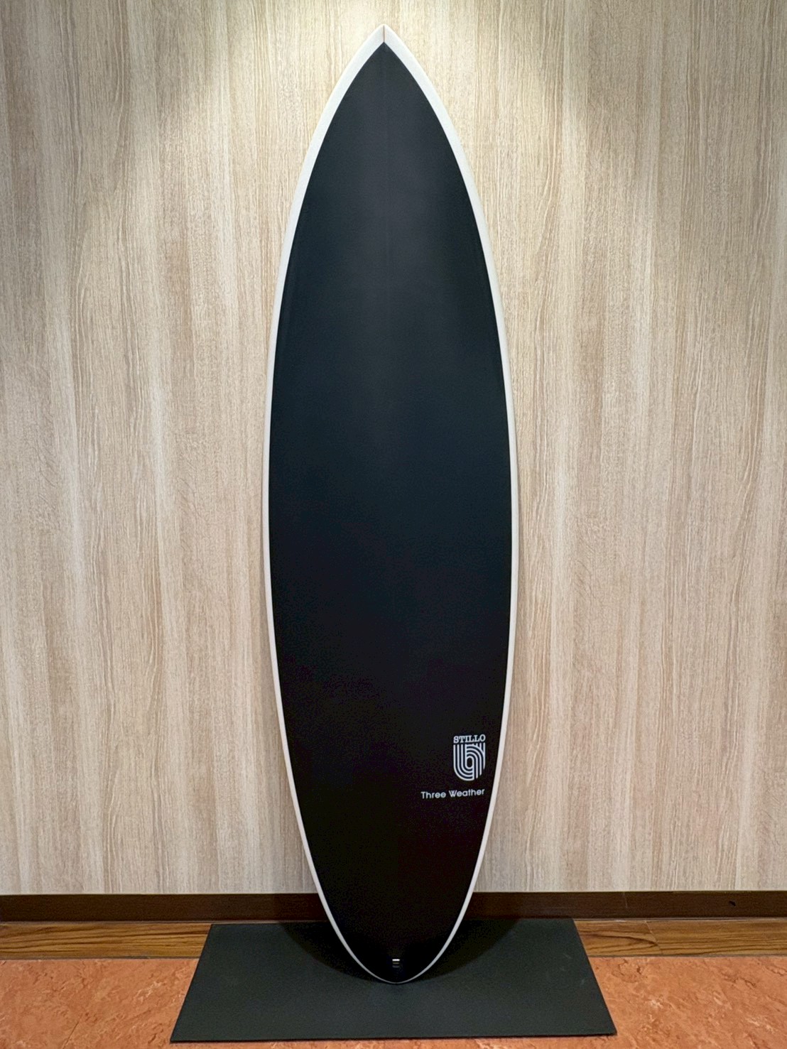 Vertical black surfboard with white rail, standing upright against a light wood panel wall on a black mat.