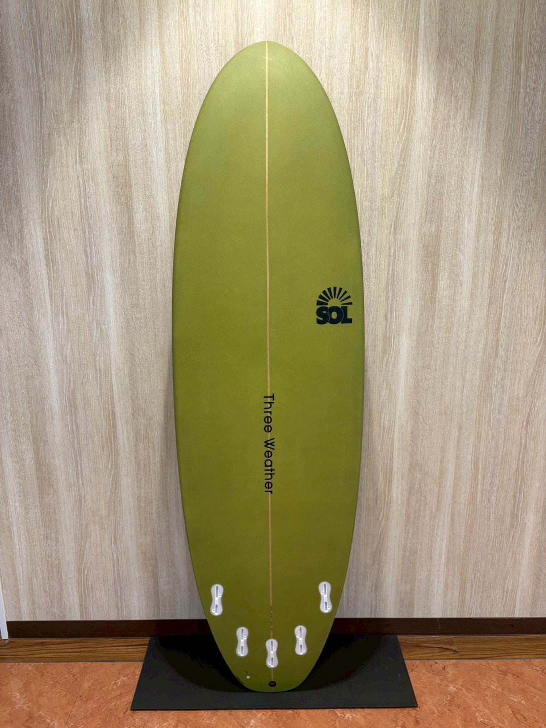 Olive-green surfboard standing upright against a wall, with a vertical stringer and six white fin boxes at the base, SOL logo on the right.