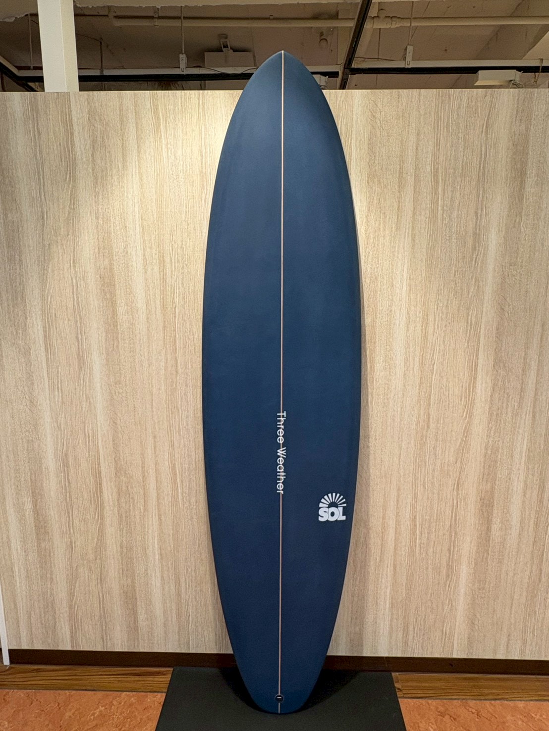 Blue surfboard on a display stand in a showroom, with a white center seam and an SOL logo near the bottom.
