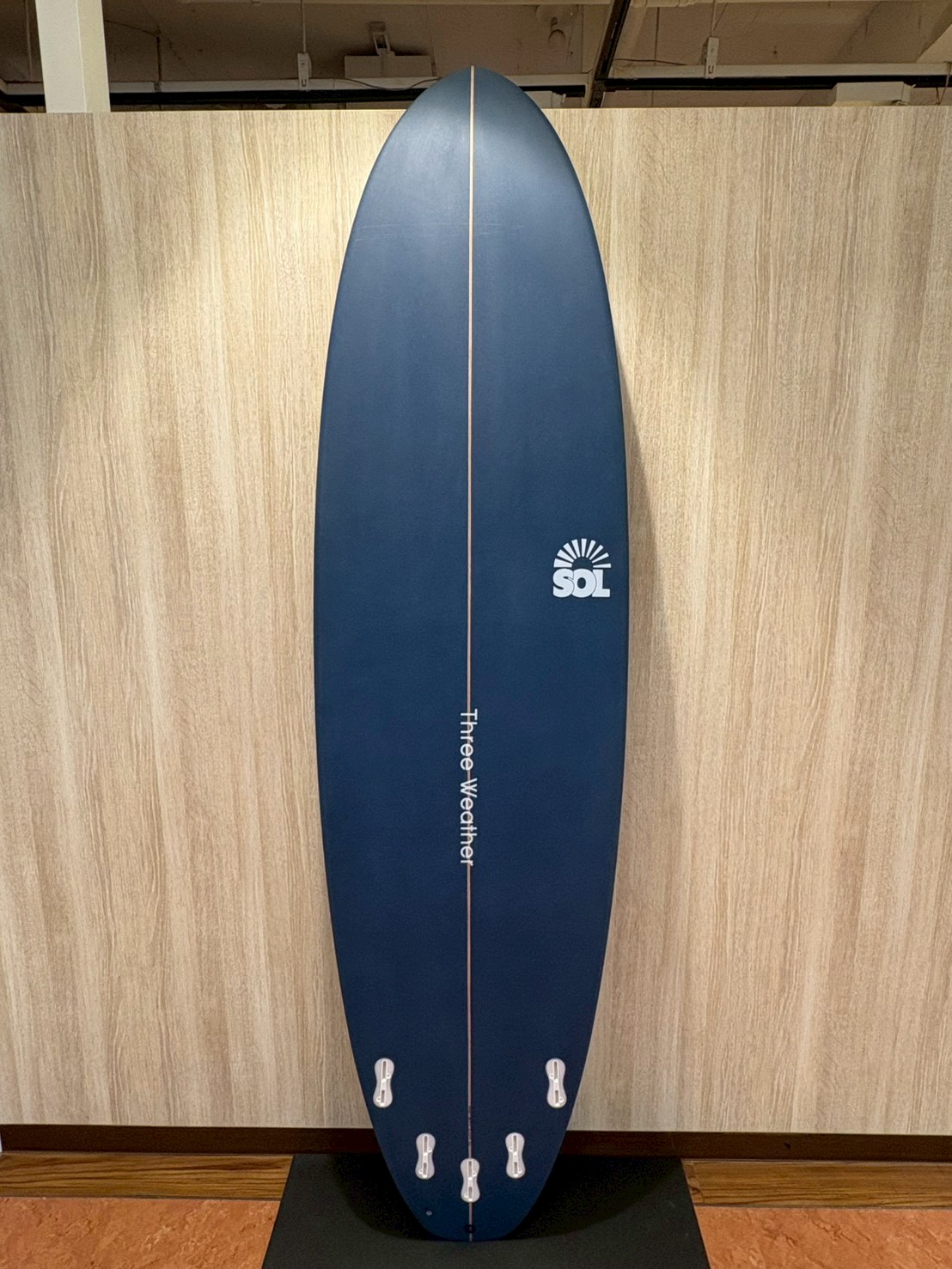 Blue surfboard standing upright against a wooden wall, with a centered stringer, five tail fins, and a SOL logo on the right side near the midsection. The vertical text along the stringer reads 'Three Weather'.