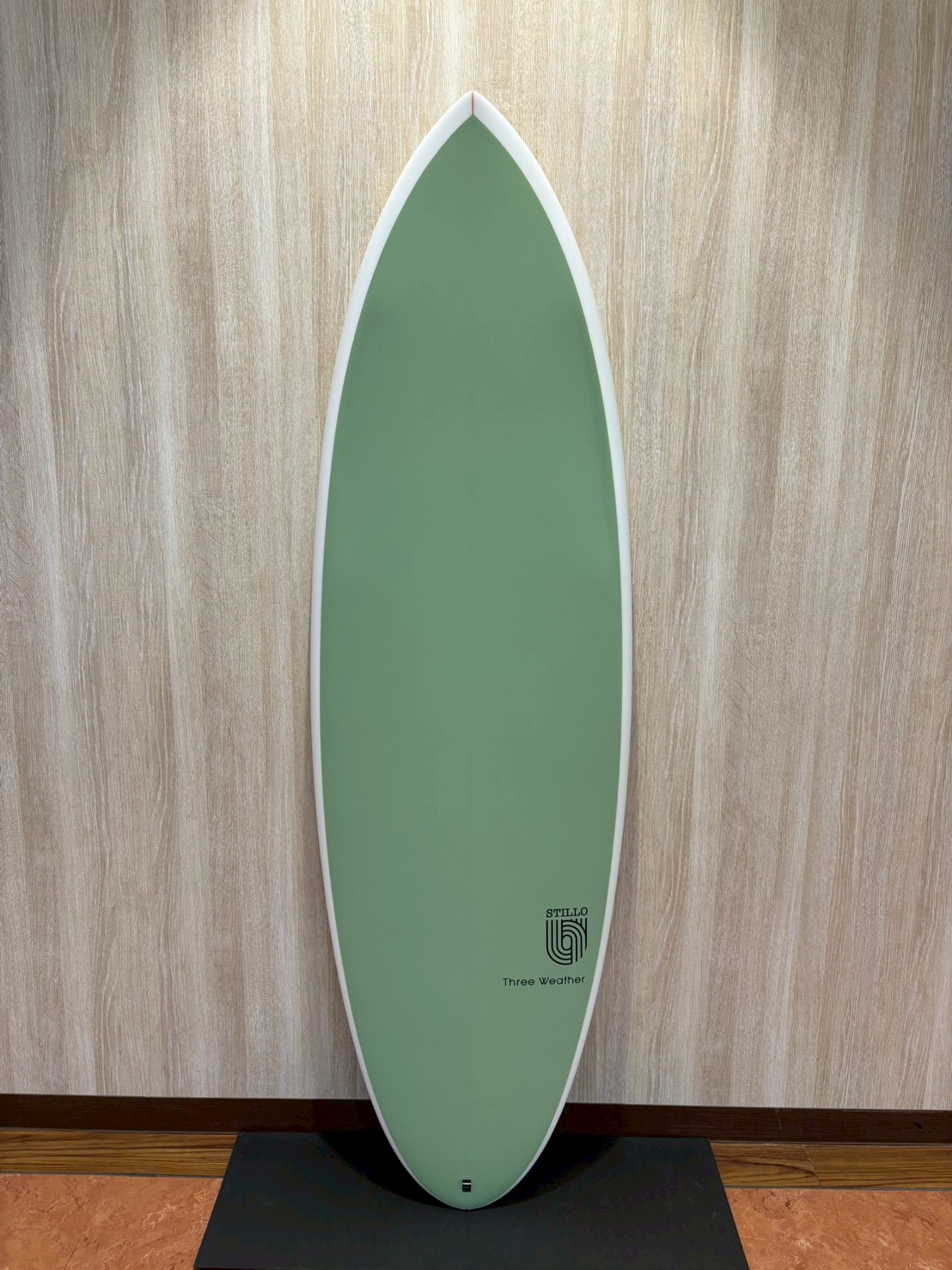 Green surfboard standing upright on a display pedestal indoors, with white edge and small logo near the bottom reading 'Three Weather'