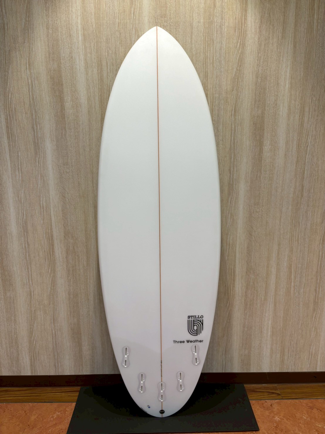 White surfboard on a display stand against a wood-textured wall, featuring an orange center stringer and six fin boxes at the tail.