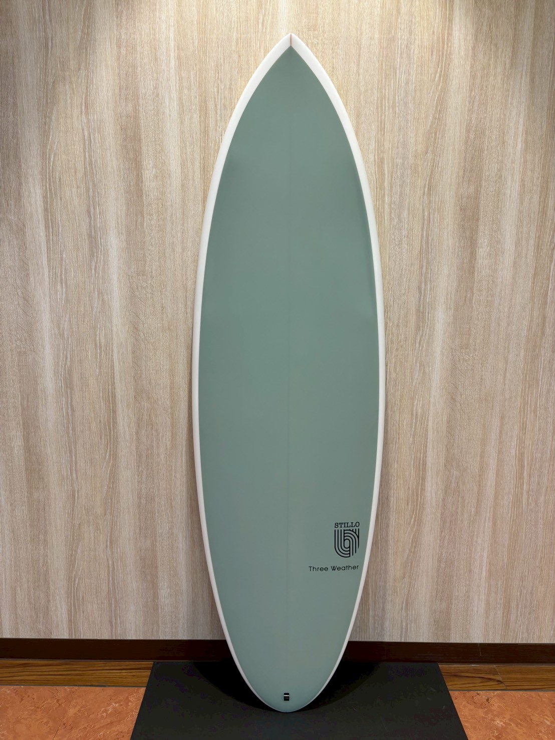 Light teal surfboard standing upright against a wooden wall with a white edge and small logo near the bottom that reads 'STILLO' and 'Three Weather'