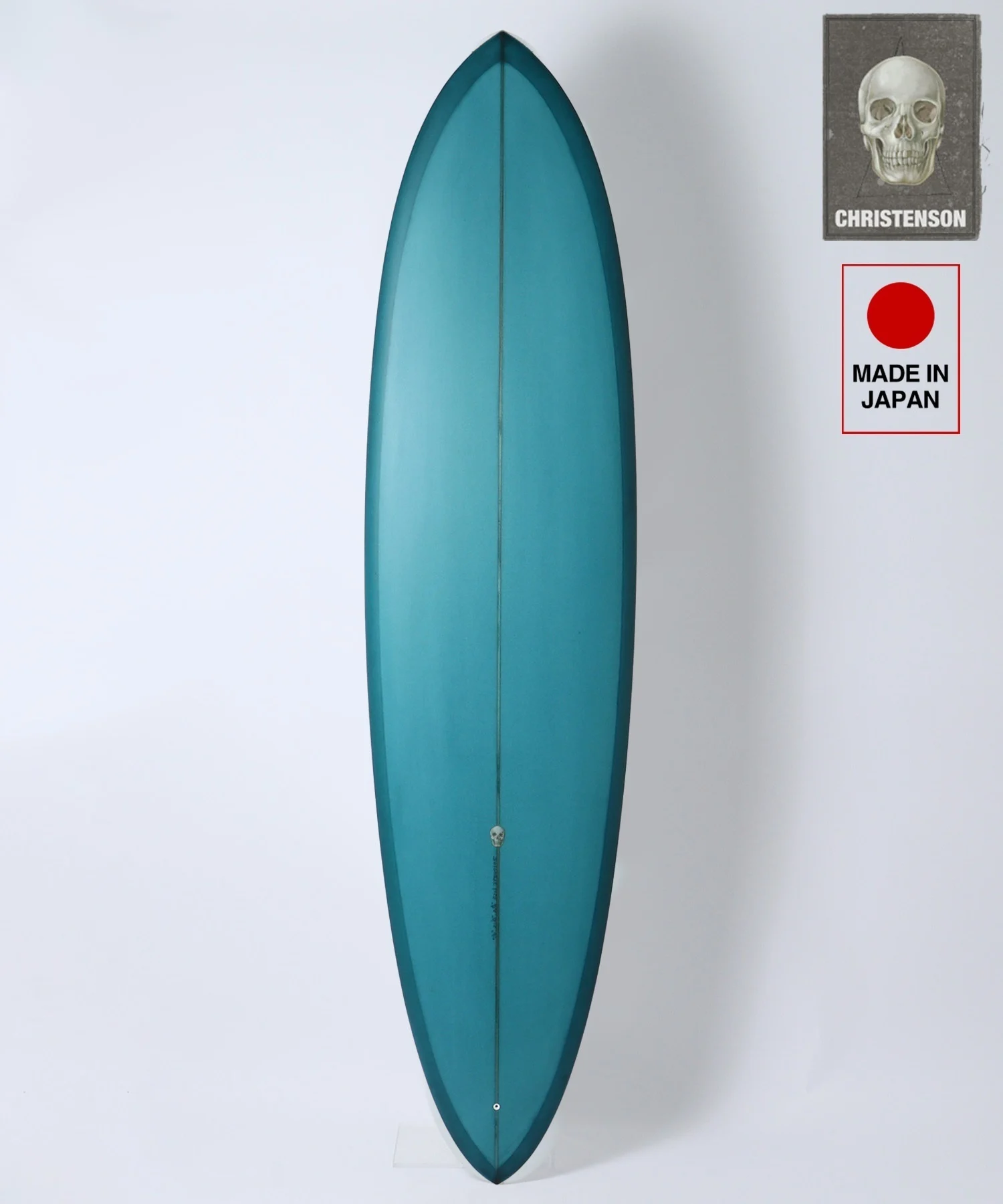 Vertical teal surfboard standing against a white wall, with two stickers on the upper right (skull 'Christenson' badge and 'Made in Japan' label).