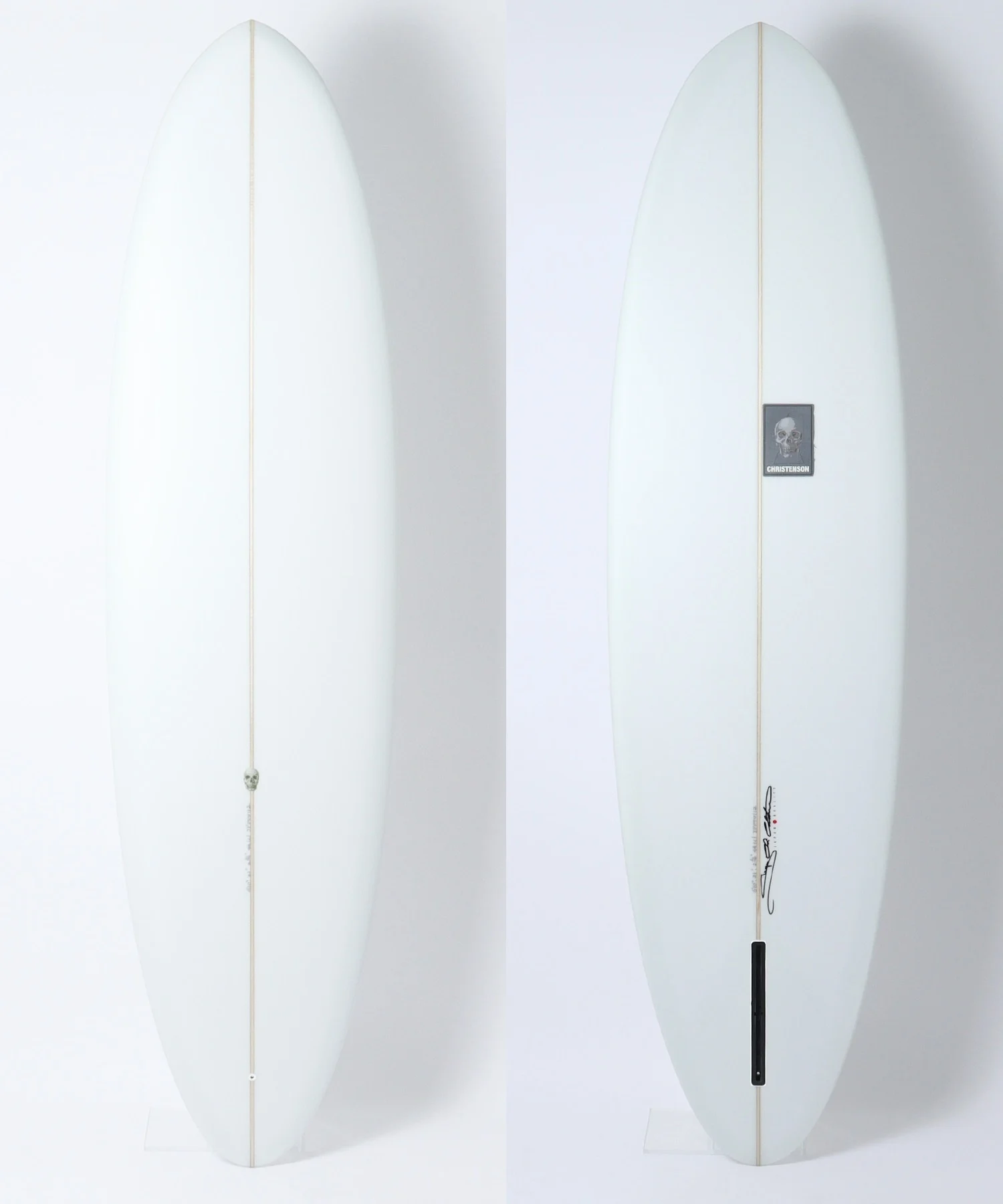 Two white surfboards standing upright side by side against a white backdrop; left board has a small skull charm on the cord running down its fin, right board shows a skull sticker and signature near the bottom.
