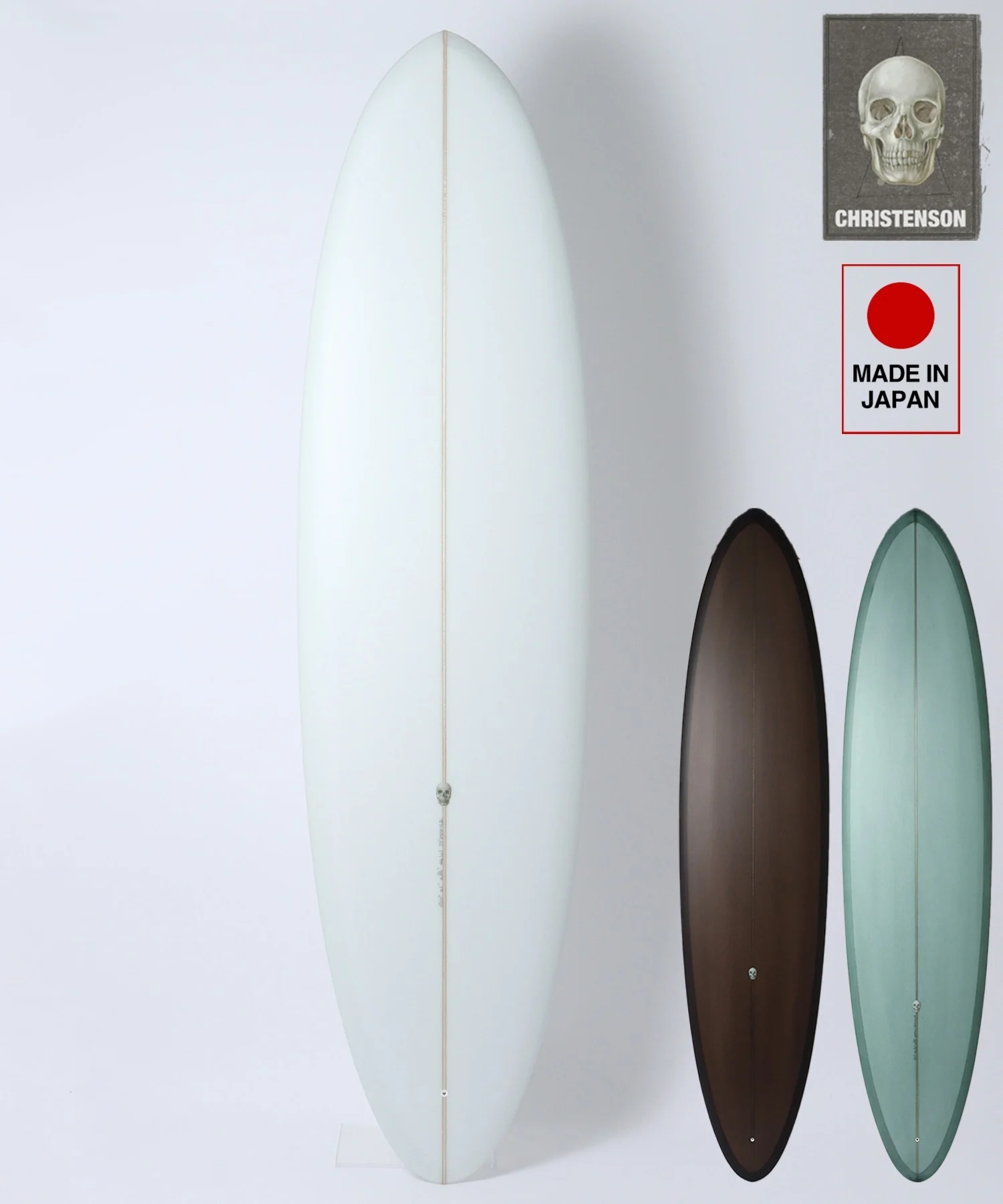 Tall pale blue surfboard standing upright against a white wall with three smaller brown, green, and black boards beside it; brand tag visible on top right.