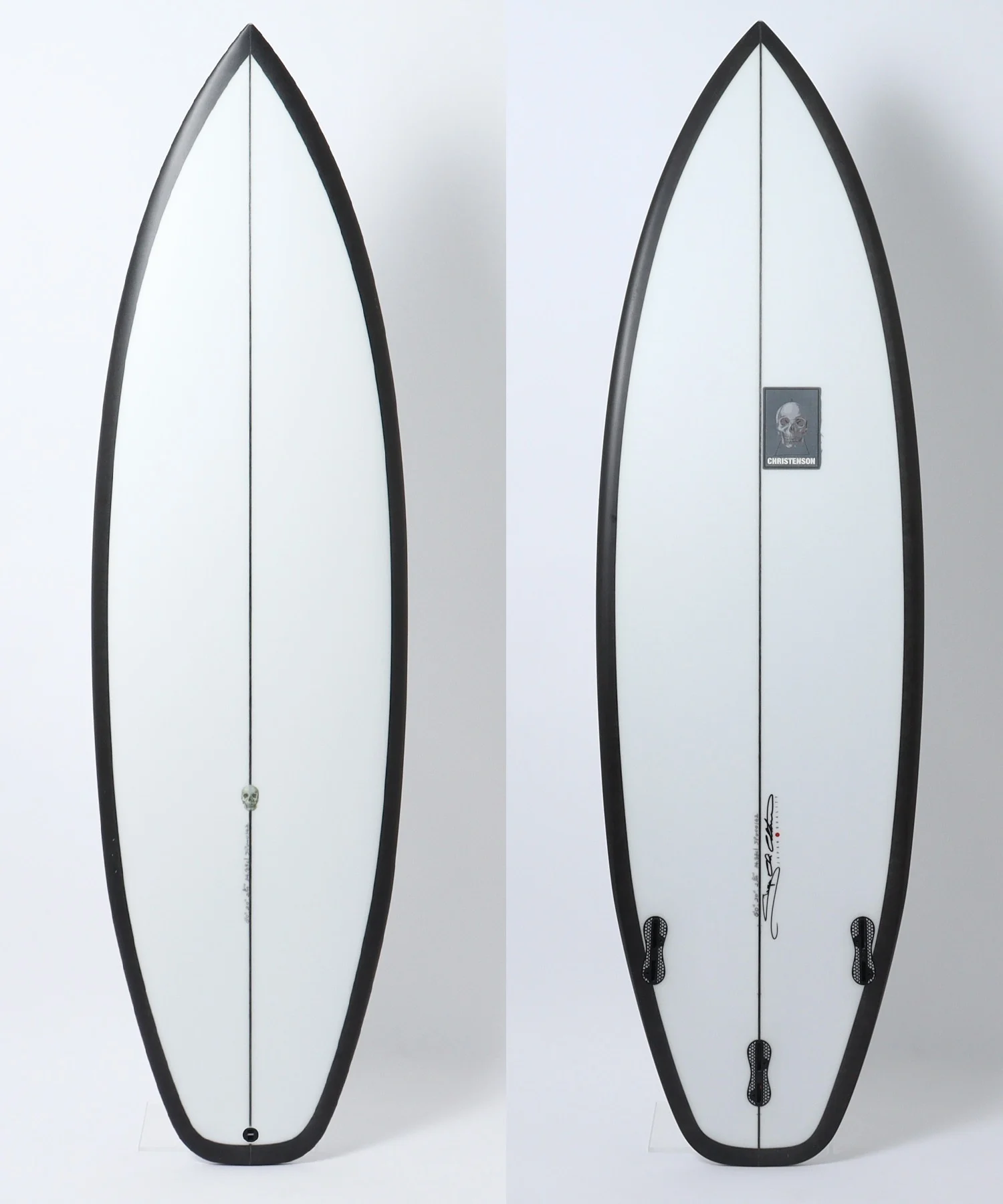 Two white surfboards shown side by side, highlighting the tail and deck details with black rails and a central stringer; left side features a skull sticker near the tail, right side shows signature and logo patch.